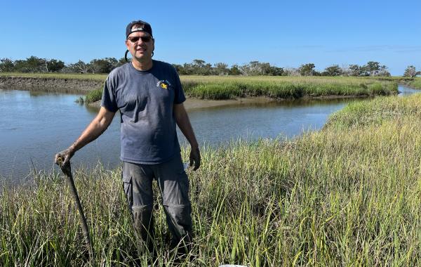 A man in a blue shirt holds a shovel in a salt marsh. 