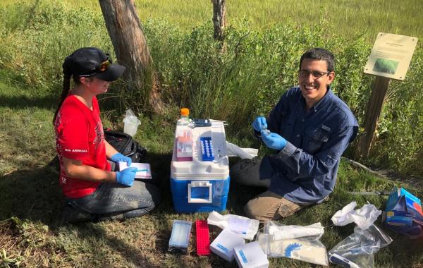 Two people sitting on a ground with a cooler and scientific equipment (including sample vials) between them. 