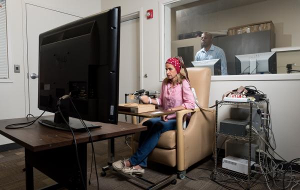 A person seated in a beige chair using a computer setup with multiple cables and devices, facing a large monitor in a testing or research room, with another individual visible through a window in an adjacent control room.