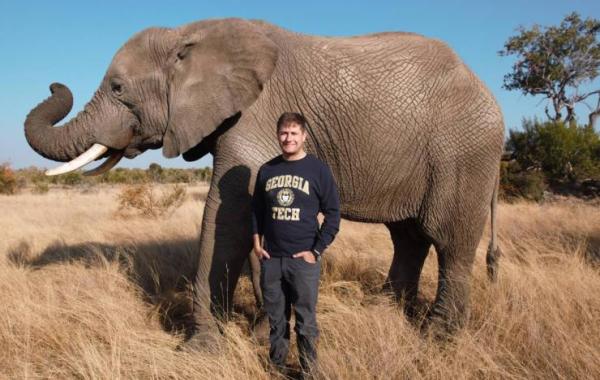 Andrew Schulz standing in front of an elephant.