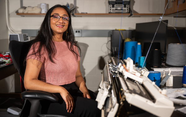 A woman wearing glasses and short sleeve pink sweater sit nexts to a commercial knitting machine.