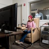 Wheaton.jpg A person seated in a beige chair using a computer setup with multiple cables and devices, facing a large monitor in a testing or research room, with another individual visible through a window in an adjacent control room.